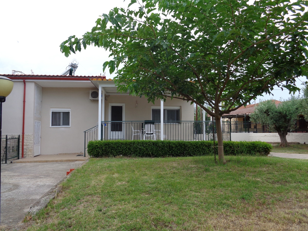 A single-story flat is surrounded by a lush garden, featuring a welcoming porch with seating. A broad path leads to the entrance, and a tree provides shade to the exterior. The area is calm, emphasizing the home's private setting.
