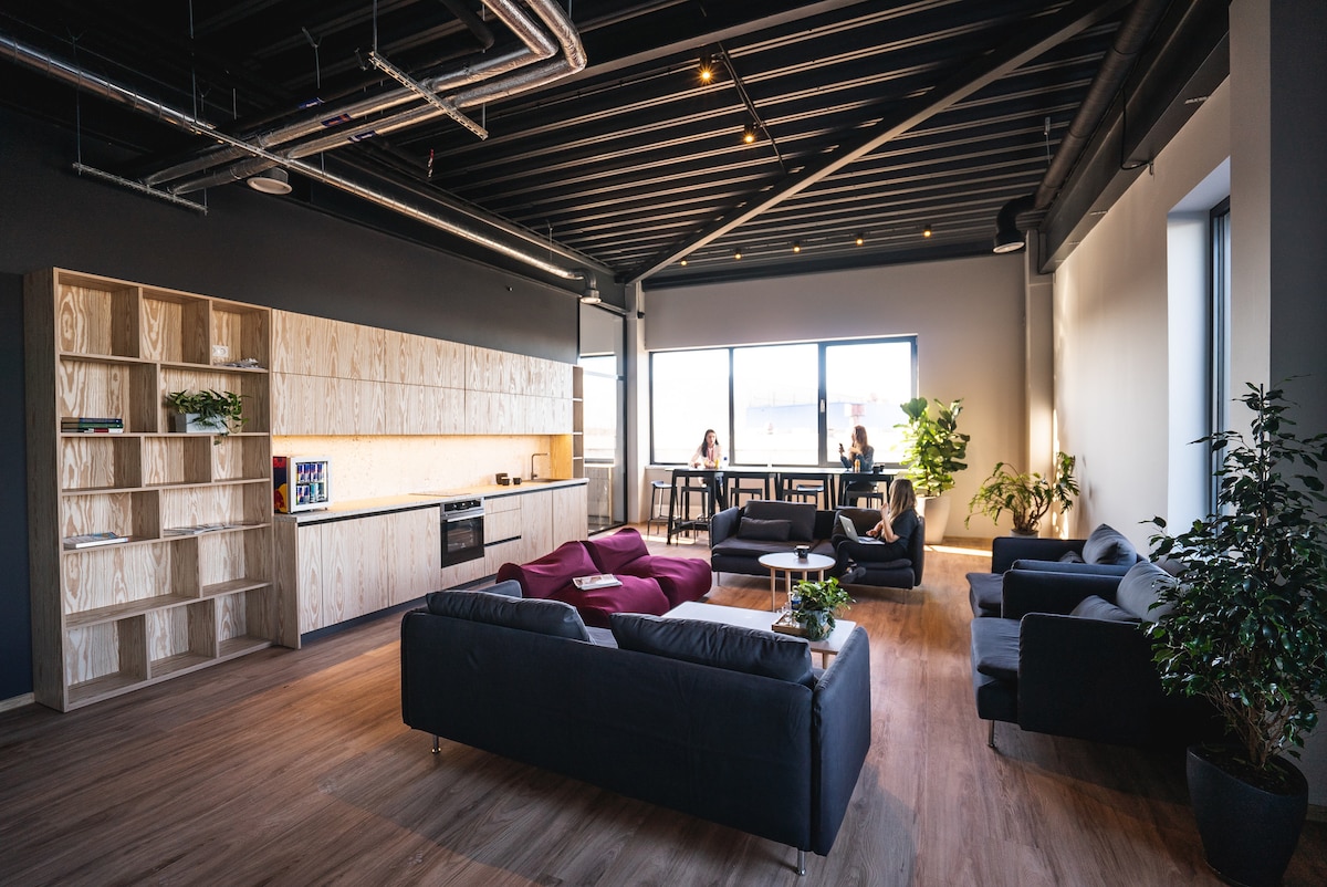 A spacious common area features sleek black sofas and vibrant red armchairs surrounding a central coffee table. A modern kitchen with wooden cabinetry and appliances is visible in the background. Large windows allow ample natural light, enhancing the open atmosphere filled with plants.