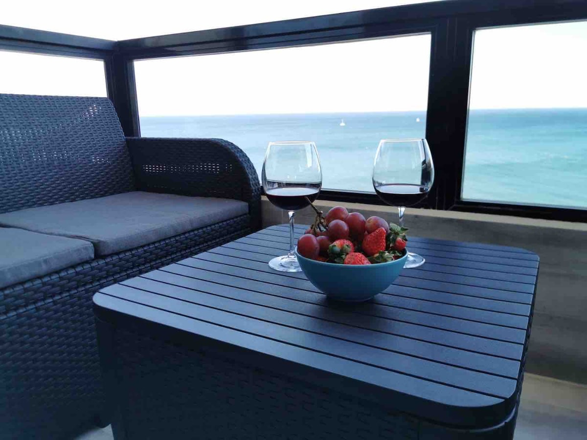 A terrace table is set with a bowl of strawberries and cherries, accompanied by two glasses of red wine. A comfortable sofa is visible in the background, framed by expansive windows revealing views of the serene Mediterranean Sea.