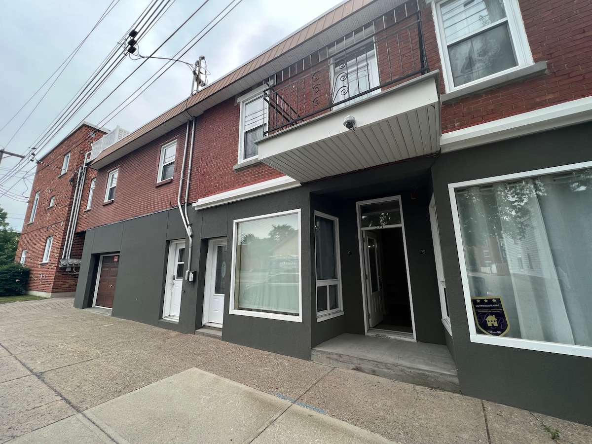 The exterior of the modern loft building is visible, featuring a mix of red brick and grey walls. The entrance is framed by several large windows, and the entryway is highlighted by a simple awning. Sidewalk access is indicated, with power lines overhead.