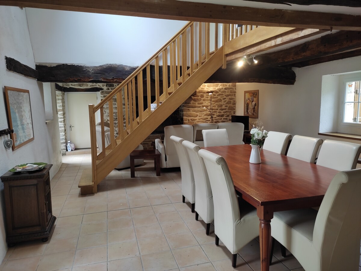 A spacious dining area features a large wooden table surrounded by white chairs, complemented by a rustic stone wall. A wooden staircase leads to an upper level, with natural light illuminating the space from multiple angles.