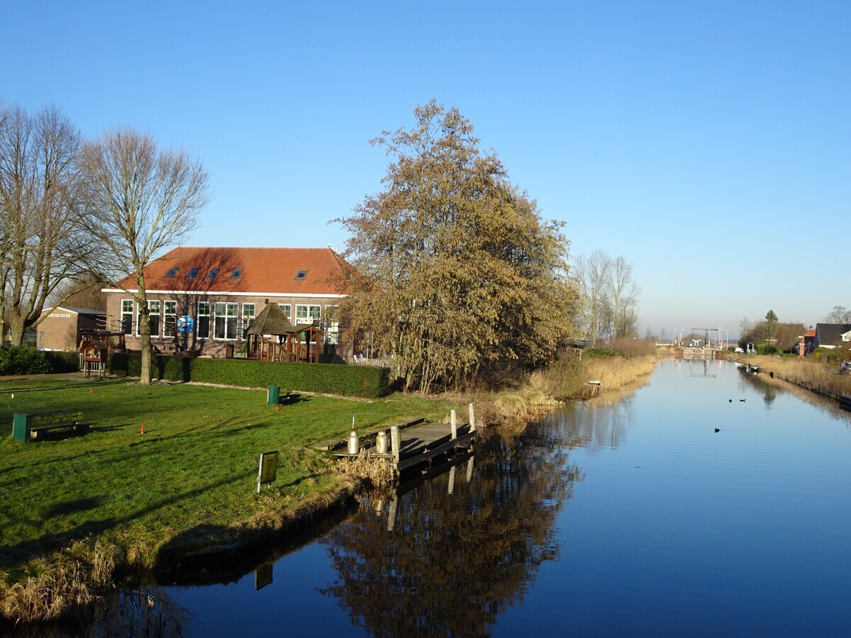 A serene view of the accommodation alongside a still waterway, reflecting the structure and surrounding greenery. The building features a red-tiled roof and large windows, with trees and open grassy areas visible nearby. Clear blue skies complement the tranquil setting.