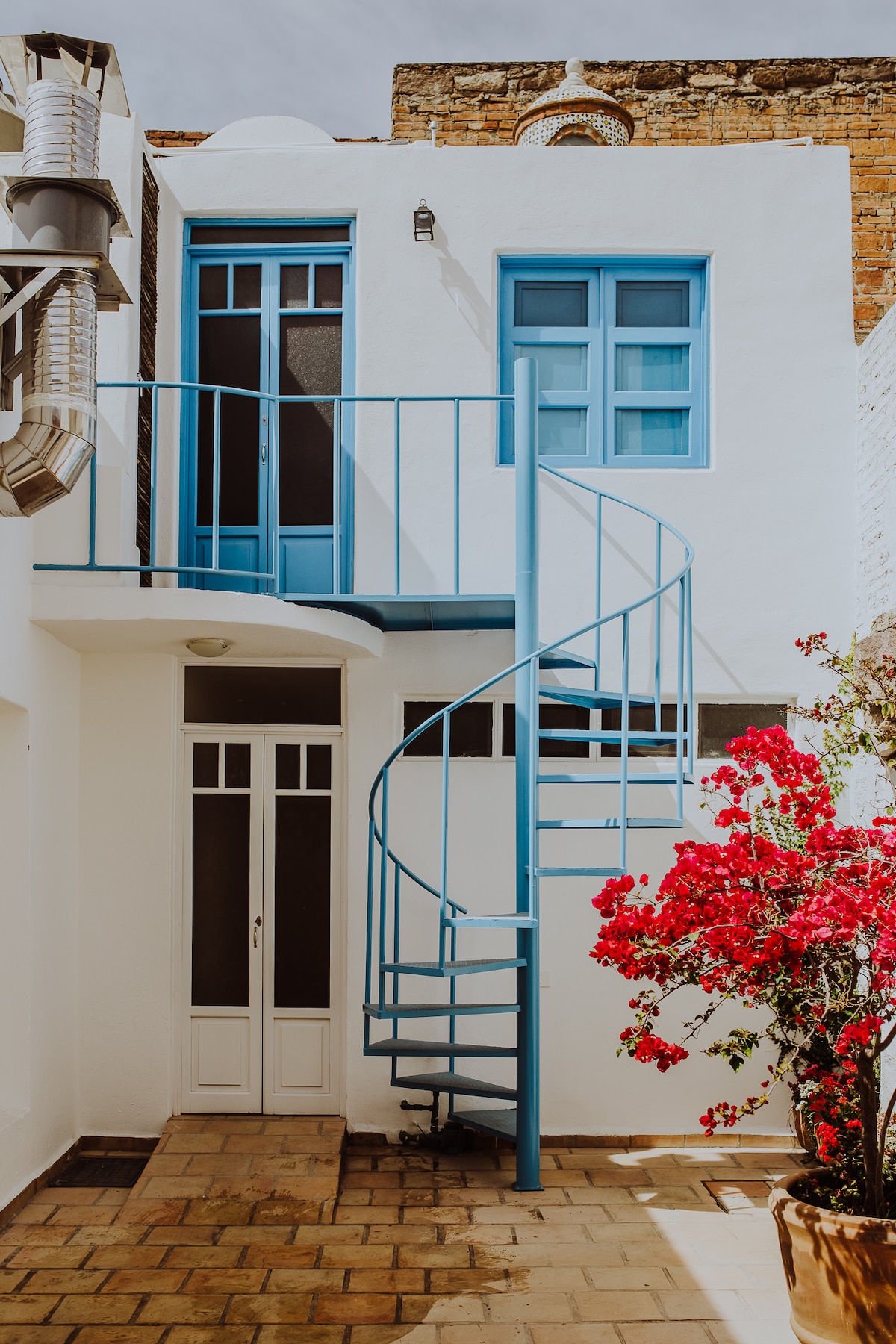 A spiral staircase in a vibrant blue color leads to an upper level, framed by white walls. A white door stands at the base, while clusters of bright bougainvillea flowers add a pop of color to the courtyard's stone floor.