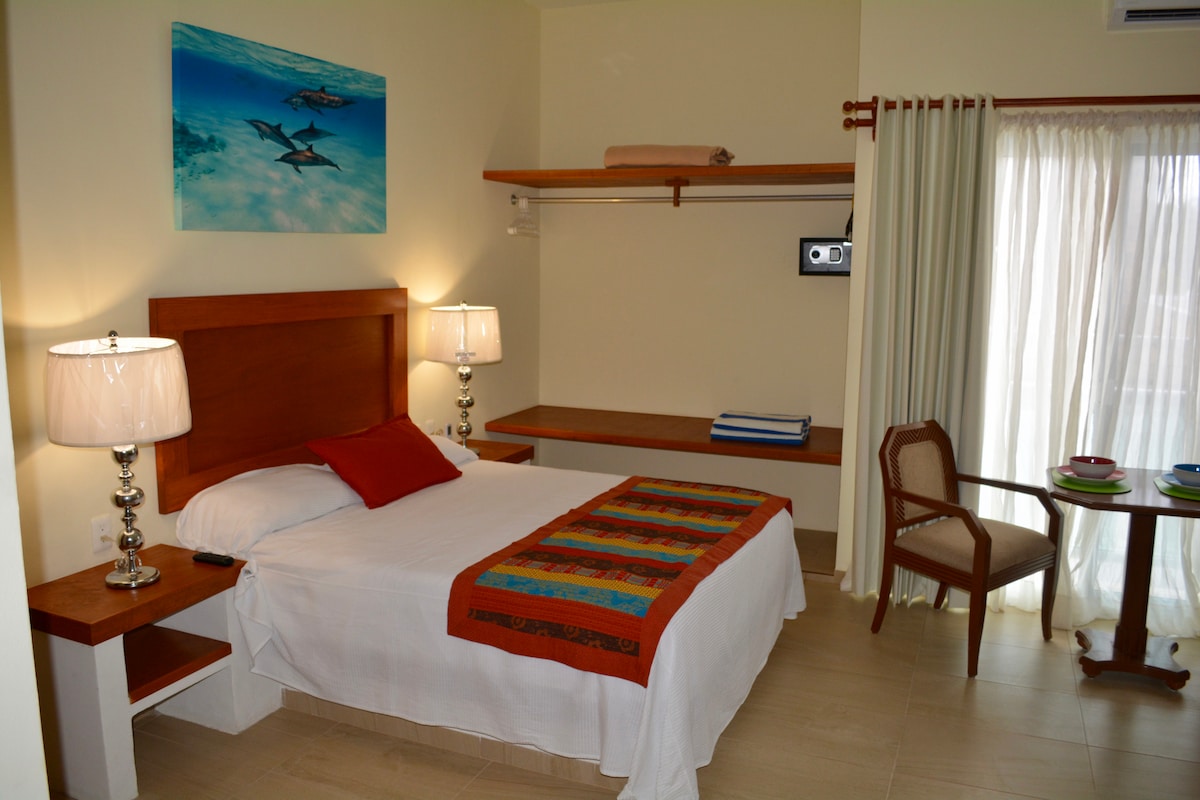 A well-appointed suite features a spacious bed with a colorful bed runner. A pair of bedside lamps flanks the headboard, and a small table is set up for dining. A bright piece of wall art depicting dolphins adds visual interest to the room.