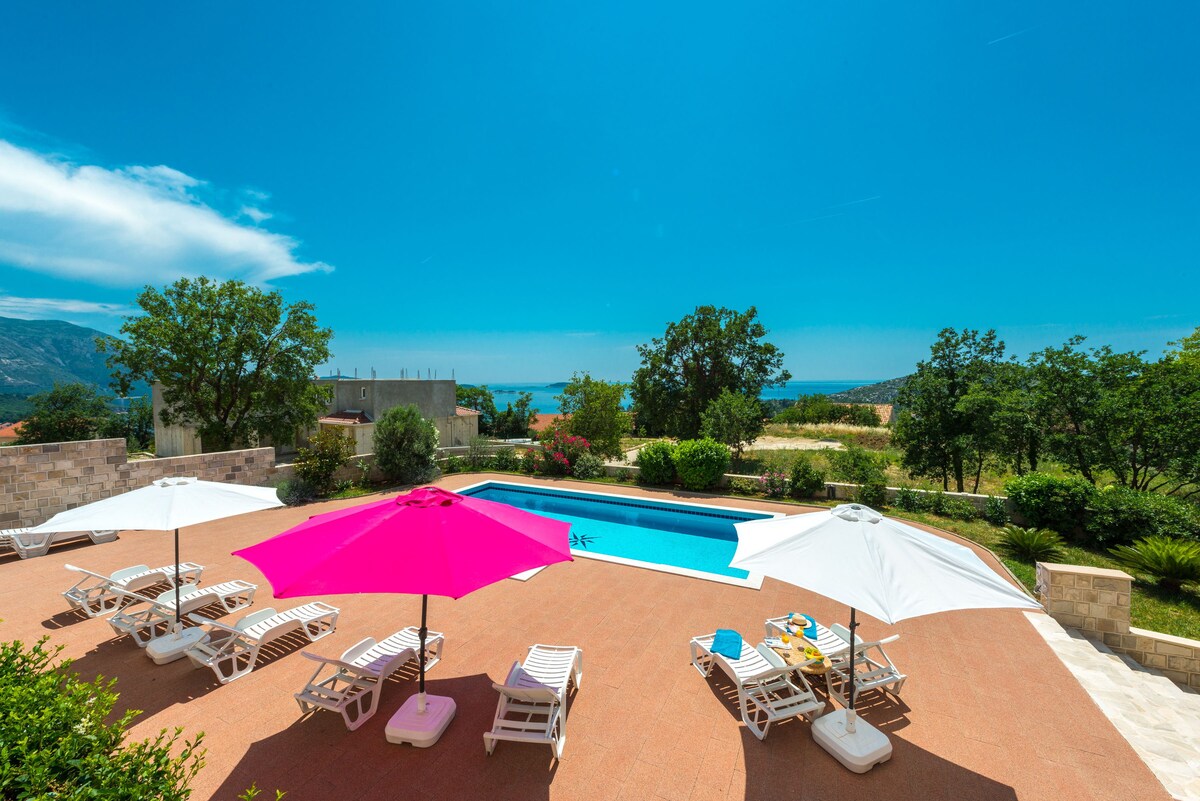 A well-maintained outdoor area features a spacious swimming pool surrounded by lounge chairs. Brightly colored umbrellas, one pink and one white, provide shade. Lush greenery and distant views of the water enhance the scenery under a clear blue sky.