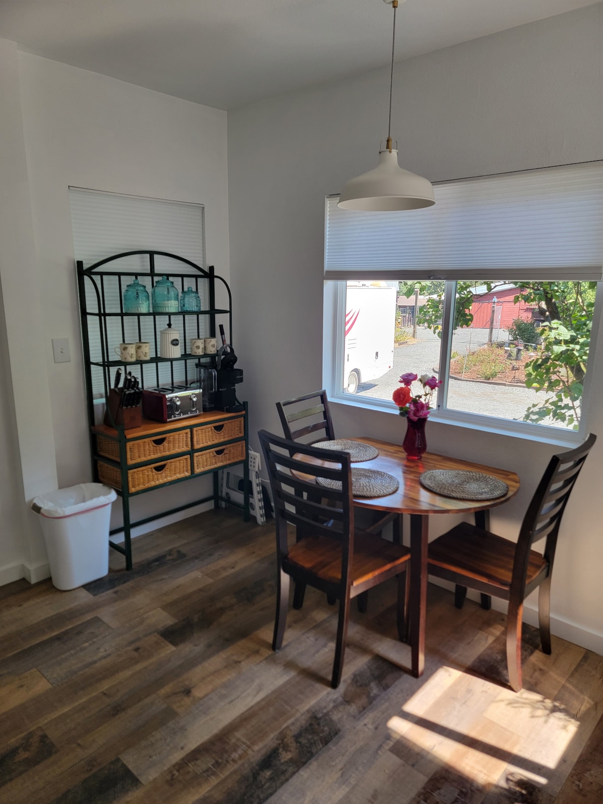 A cozy dining area features a round table surrounded by wooden chairs. A small storage unit displays kitchen essentials, while a window allows natural light to illuminate the space. A waste bin is positioned nearby, adding to the room's functionality.