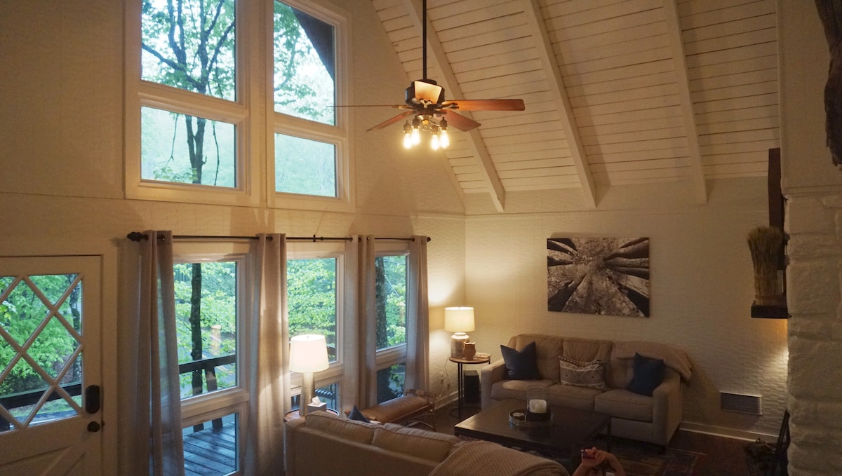 A spacious living room features tall windows that allow natural light to fill the space, showcasing views of the surrounding greenery. A comfortable seating arrangement is complemented by a ceiling fan and light fixtures, creating a cozy atmosphere for relaxation. Decorative art adds visual interest.