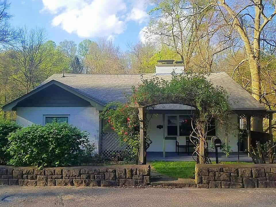 The cozy cottage is framed by lush greenery, featuring a covered front porch with seating. A stone wall lines the front, and trees are visible in the background, creating a serene natural setting. Soft light filters through the leaves, enhancing the peaceful ambiance.