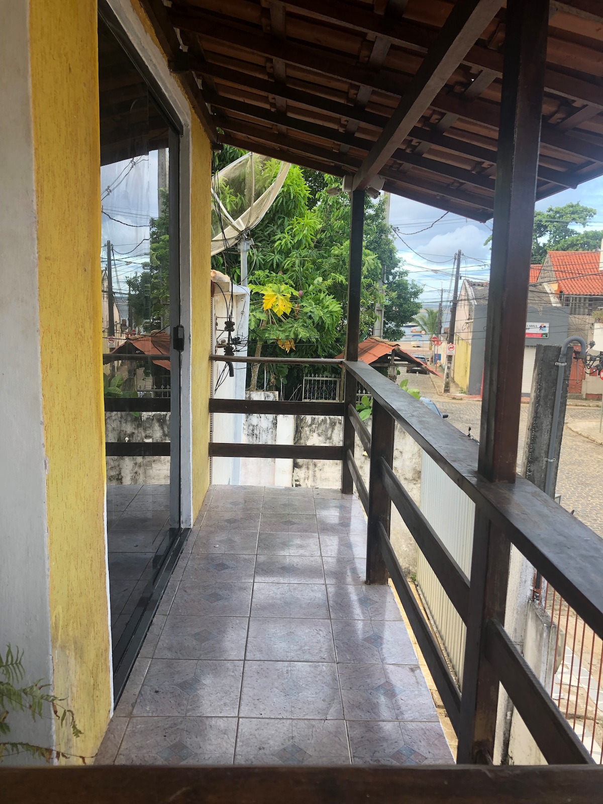 A small balcony features a covered wooden railing, providing views of the street below. The tiled floor enhances the outdoor space, while surrounding greenery can be seen in the background. Large glass doors connect the balcony to the indoor area.