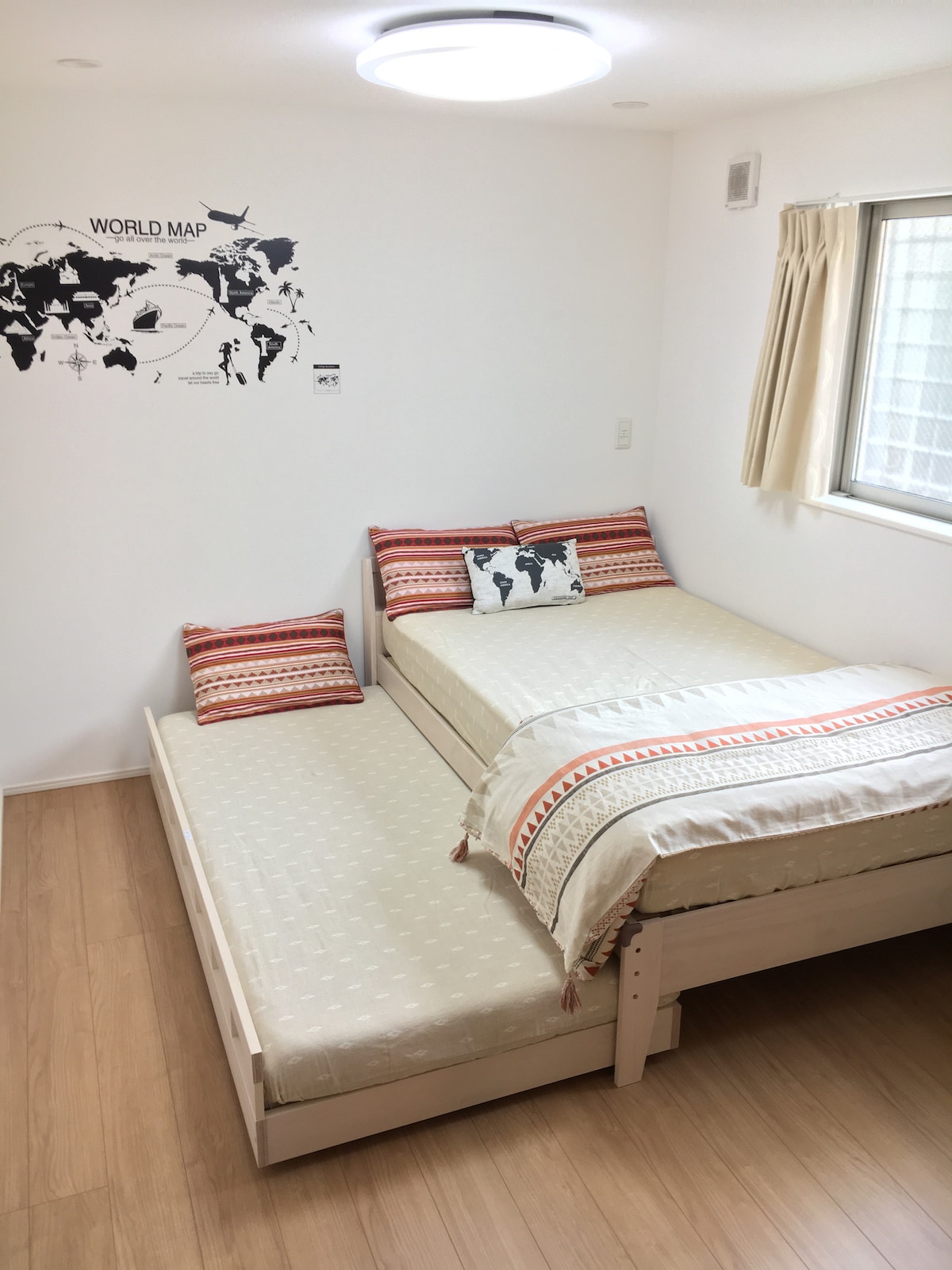 The sleeping area features a minimalist design with a spacious bed and an additional trundle bed. Decorative pillows with a world map motif are arranged on the beds. Natural light enters through a window, and light-colored walls contribute to a bright atmosphere.