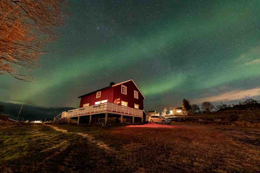 An old Norwegian house is depicted against a vibrant night sky illuminated by the northern lights. The exterior features a red facade, while the surrounding landscape includes a grassy area and visible parking. A warm glow emanates from the house's windows.