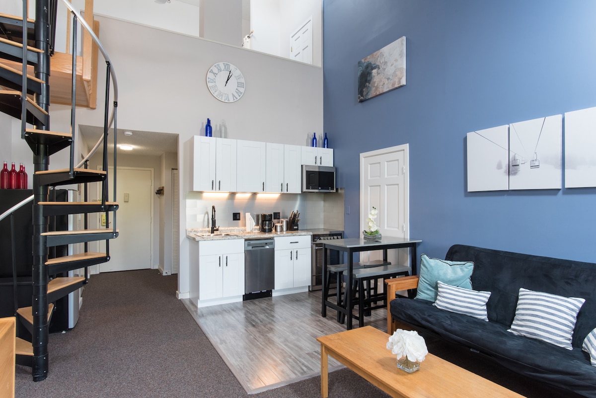 An open-concept living area is highlighted by a stylish kitchen featuring stainless steel appliances. A spiral staircase leads to the lofted bedroom. The space includes a comfortable seating area with a dark sofa and a wooden coffee table adorned with a decorative vase.