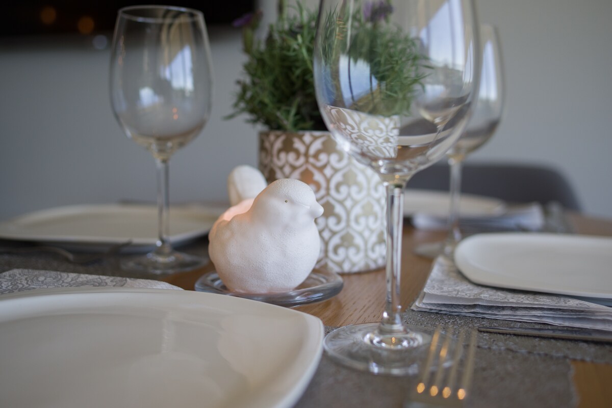 A dining table is set with white plates, elegant glassware, and silver utensils. A decorative bird figurine is placed on a glass dish, surrounded by fresh greenery in a patterned planter. Soft lighting creates a calming ambiance.