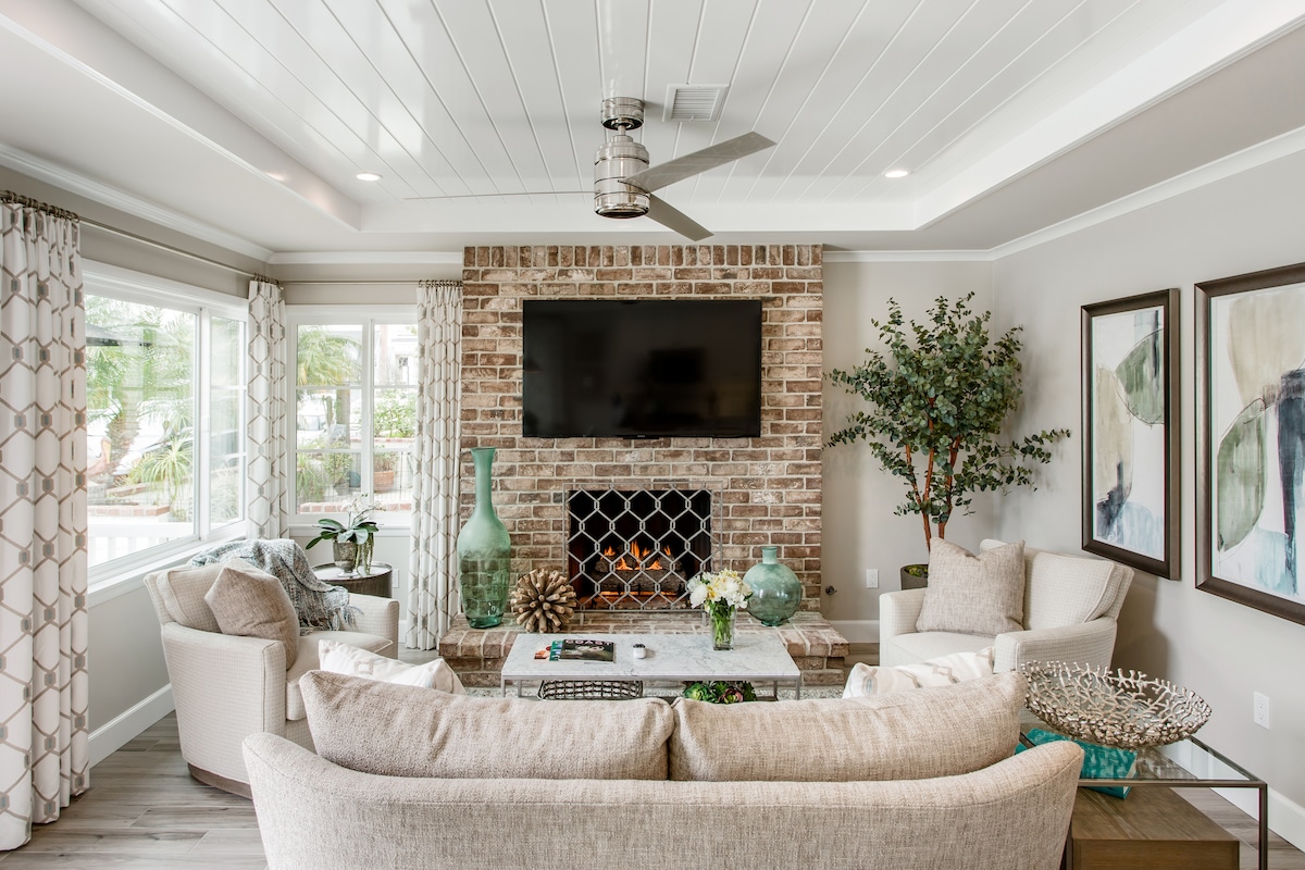A welcoming living area features a comfortable arrangement of light-toned sofas around a central coffee table. A brick fireplace adorned with decorative vases is complemented by large windows, allowing natural light to fill the space. Neutral artwork enhances the calming atmosphere.