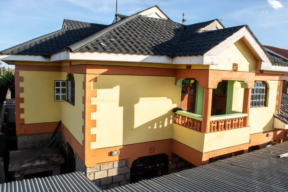 A multi-story house is set against a clear sky, featuring yellow outer walls and a patterned roof. A balcony with decorative railings is visible, along with multiple windows. The structure is framed by surrounding rooftops and greenery in the background.