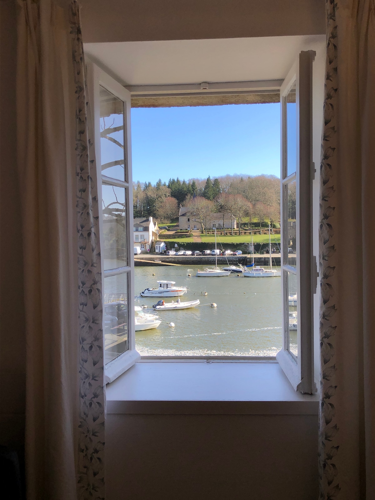 A serene view from a window showcases the tranquil harbor filled with small boats. Lush greenery lines the waterfront, with buildings nestled along the shore. Soft light filters through the open window, illuminating the curtains with a floral design.