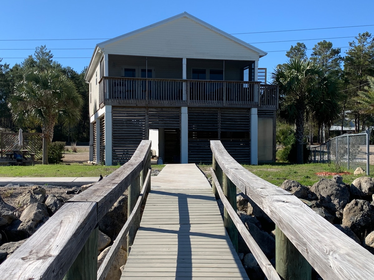 A wooden dock leads to a two-story modular home with a spacious front porch and wooden railings. The house is situated on stilts, surrounded by green grass and palm trees, providing a relaxed coastal ambiance.