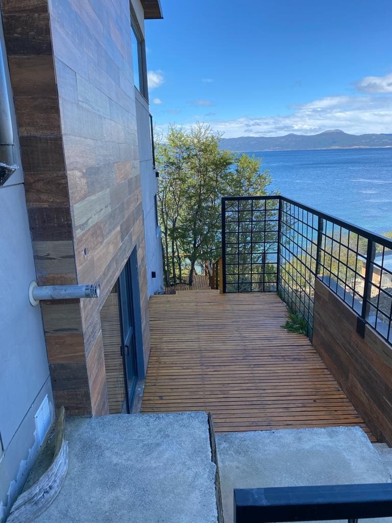 A wooden staircase leads down towards a scenic waterfront, with trees lining the path. The exterior features natural wood paneling, and glimpses of the tranquil water can be seen in the background, framed by distant mountains under a clear blue sky.