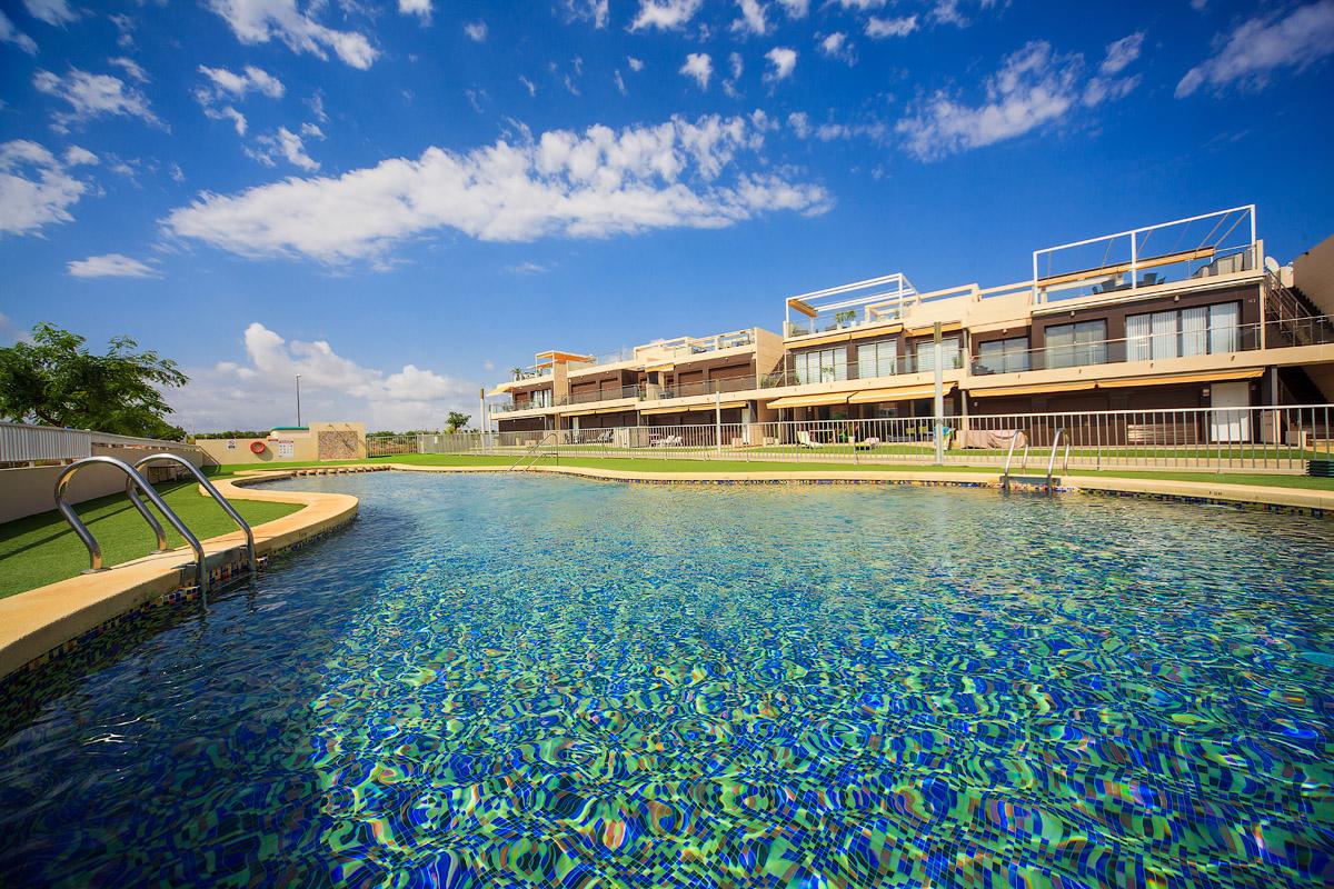 A serene swimming pool is surrounded by manicured grass and modern buildings. The water's surface reflects the clear blue sky and fluffy white clouds. A gentle shimmer enhances the inviting atmosphere of the pool area, complemented by a low wall for safety.