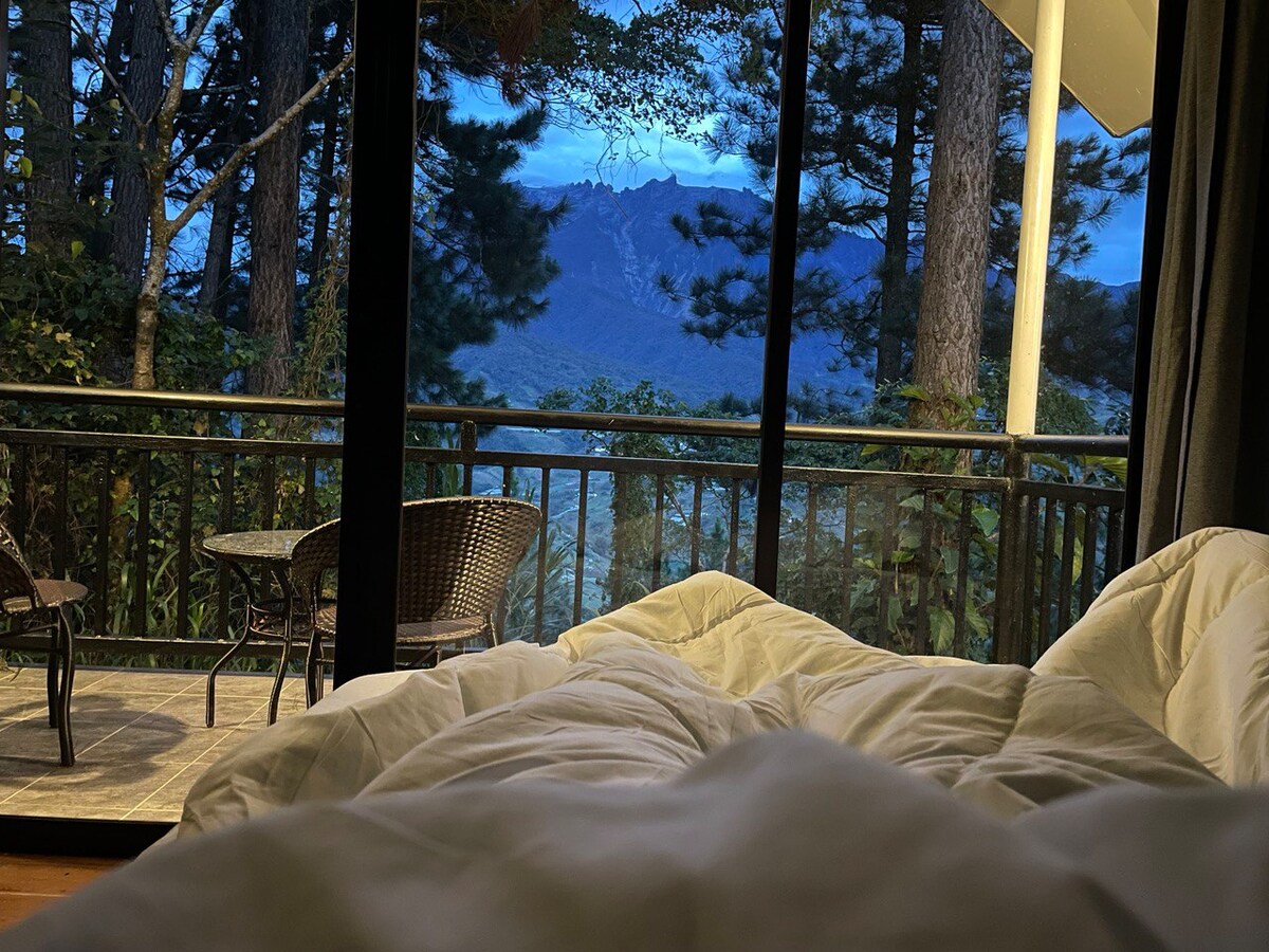 A spacious bed is covered with white bedding, positioned in front of large glass doors. The view reveals lush trees and distant mountains, while a small outdoor seating area with two wicker chairs and a circular table is visible on the porch.