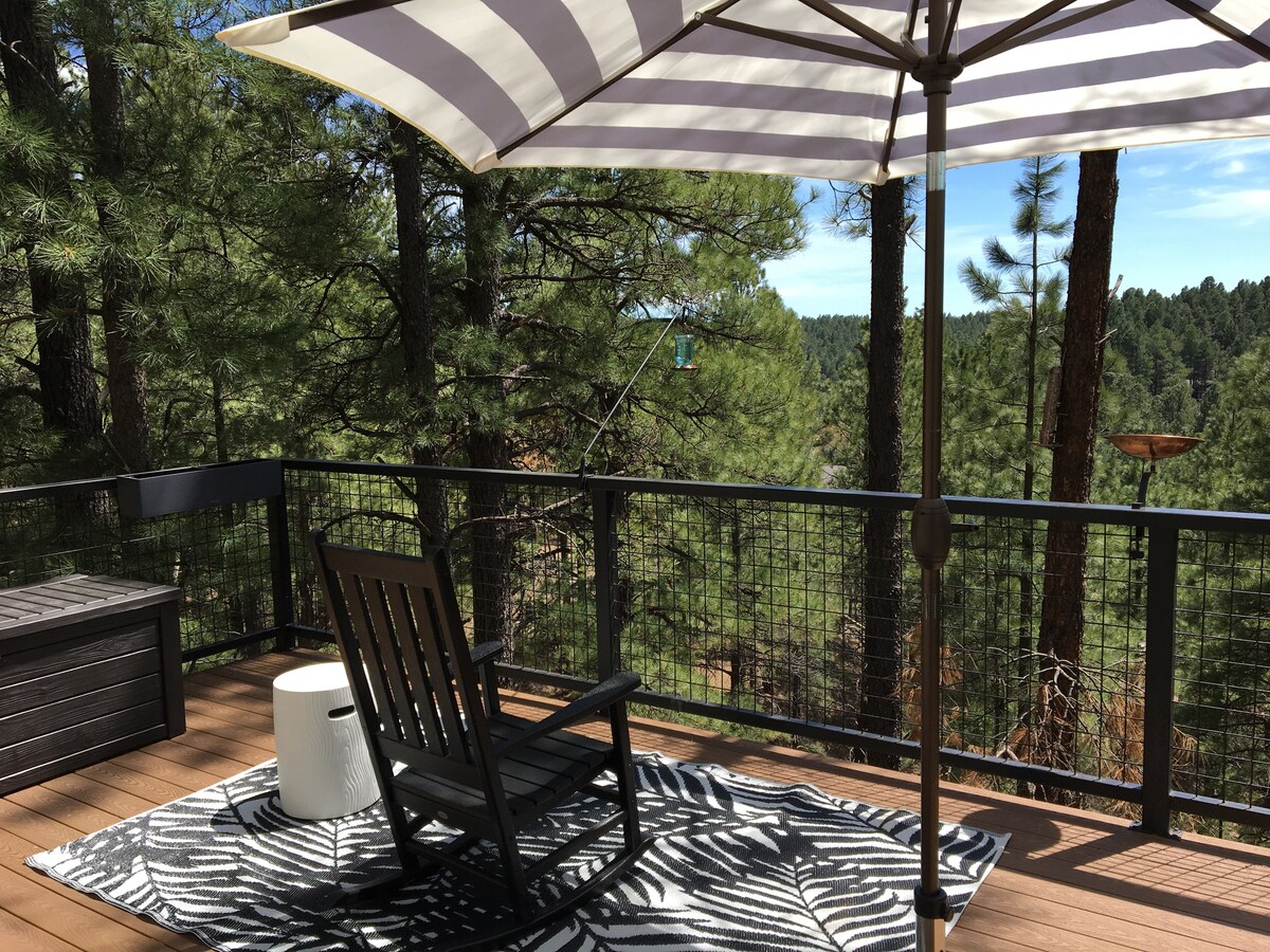 A wooden deck features a striped umbrella providing shade over a black rocking chair. An outdoor rug displays a leaf pattern, and a small white table complements the seating area. Surrounding trees enhance the serene forest setting.