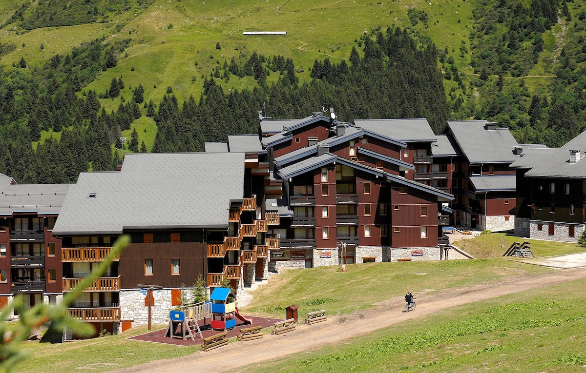 The image captures a cluster of chalet-style buildings nestled in a lush green landscape. The structures exhibit wooden balconies and stone foundations, set against a backdrop of rolling hills and forested areas, contributing to a serene mountain retreat.
