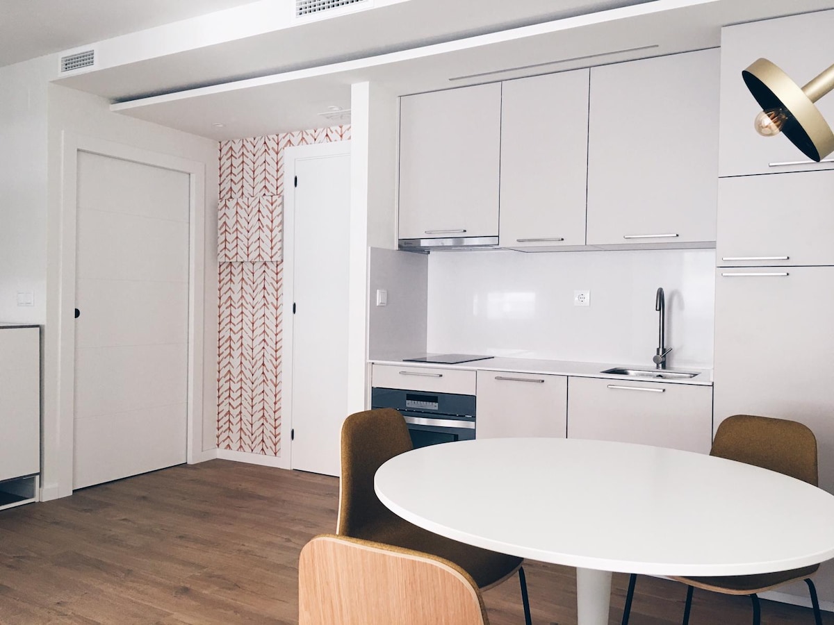 A modern kitchen area features sleek cabinetry in light tones, complemented by a circular dining table with wooden and upholstered chairs. A compact sink is situated near the kitchen appliances. The light wood flooring enhances the room's bright and clean design.