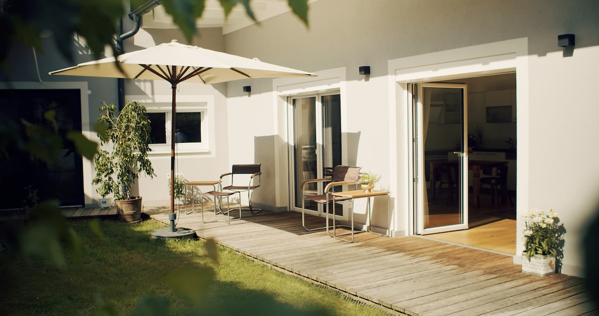 An outdoor terrace features a large umbrella providing shade over two chairs and a small table. Lush greenery surrounds the space, while glass doors lead into the interior of the home, creating an inviting transition between indoor and outdoor areas.