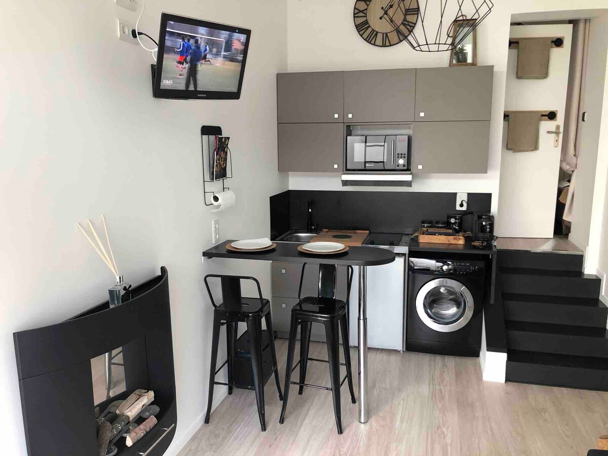 A compact kitchen area is presented, featuring modern cabinetry in neutral tones. Essential appliances, including a microwave and dishwasher, are visible. A small dining table with two chairs is positioned nearby, complemented by a decorative wall-mounted television and stylish lighting elements.