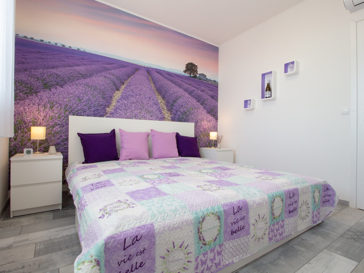 A spacious bedroom features a large bed with decorative pillows, a quilt showcasing purple tones, and bedside tables with lamps. A lavender field mural adorns the wall, creating a calming backdrop, while light enters from a nearby window covered by sheer curtains.