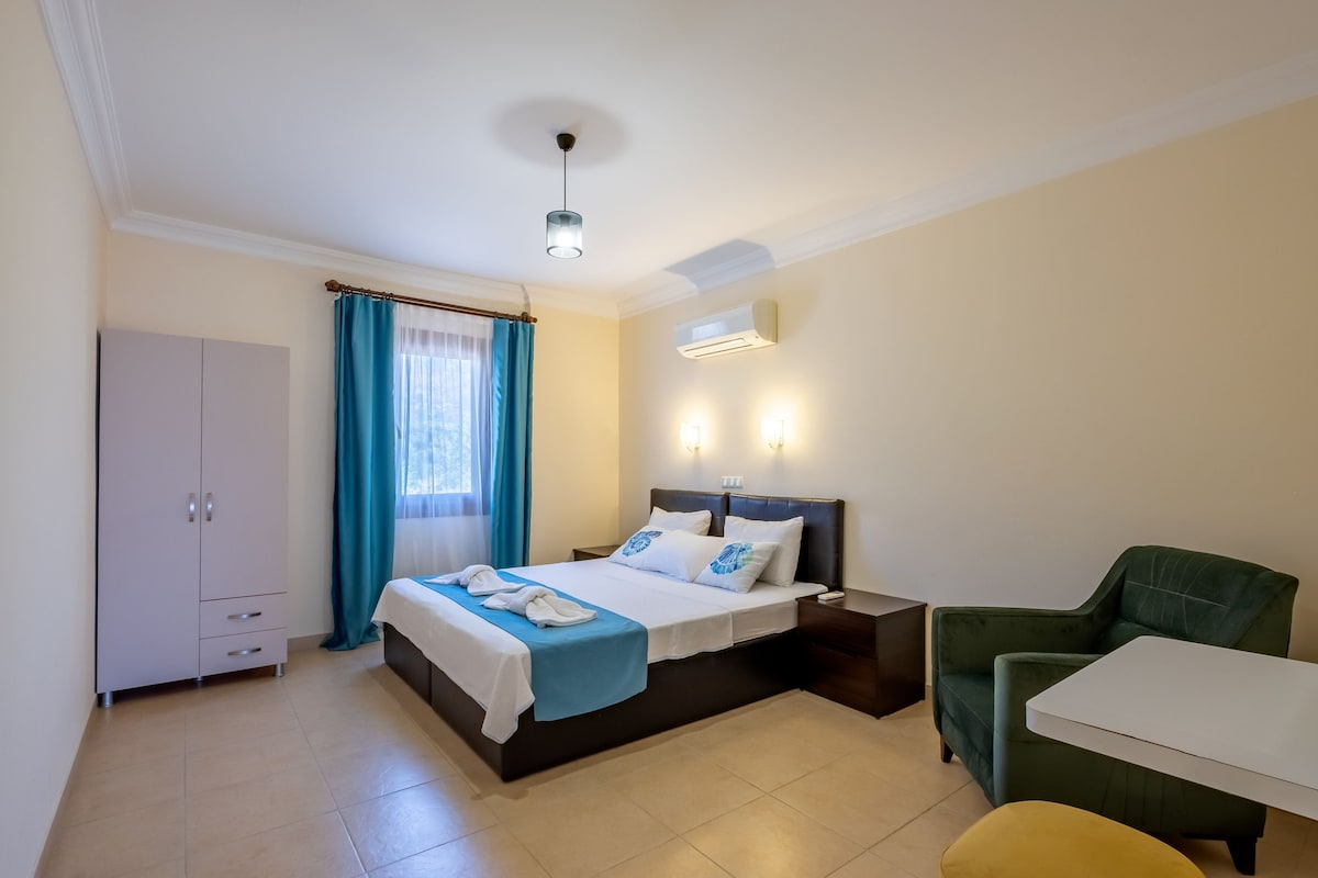 A spacious bedroom is highlighted by a large bed with crisp linens and decorative pillows. A window with sheer curtains allows natural light to enter, while a comfortable green armchair and a small table complement the furnishings. A wardrobe is positioned against the wall.