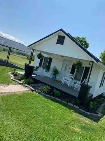 A charming white cottage with a black roof is set amid green grass and trees. A welcoming porch features two rocking chairs, complemented by flower pots. A stone pathway leads to the entrance, enhancing the home's inviting appearance.