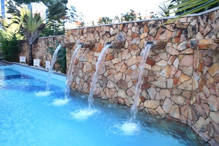 A tranquil pool area features a stone wall with four distinct water features flowing gently into the inviting blue water. Surrounding greenery adds a natural touch to the serene atmosphere, creating a relaxing retreat.