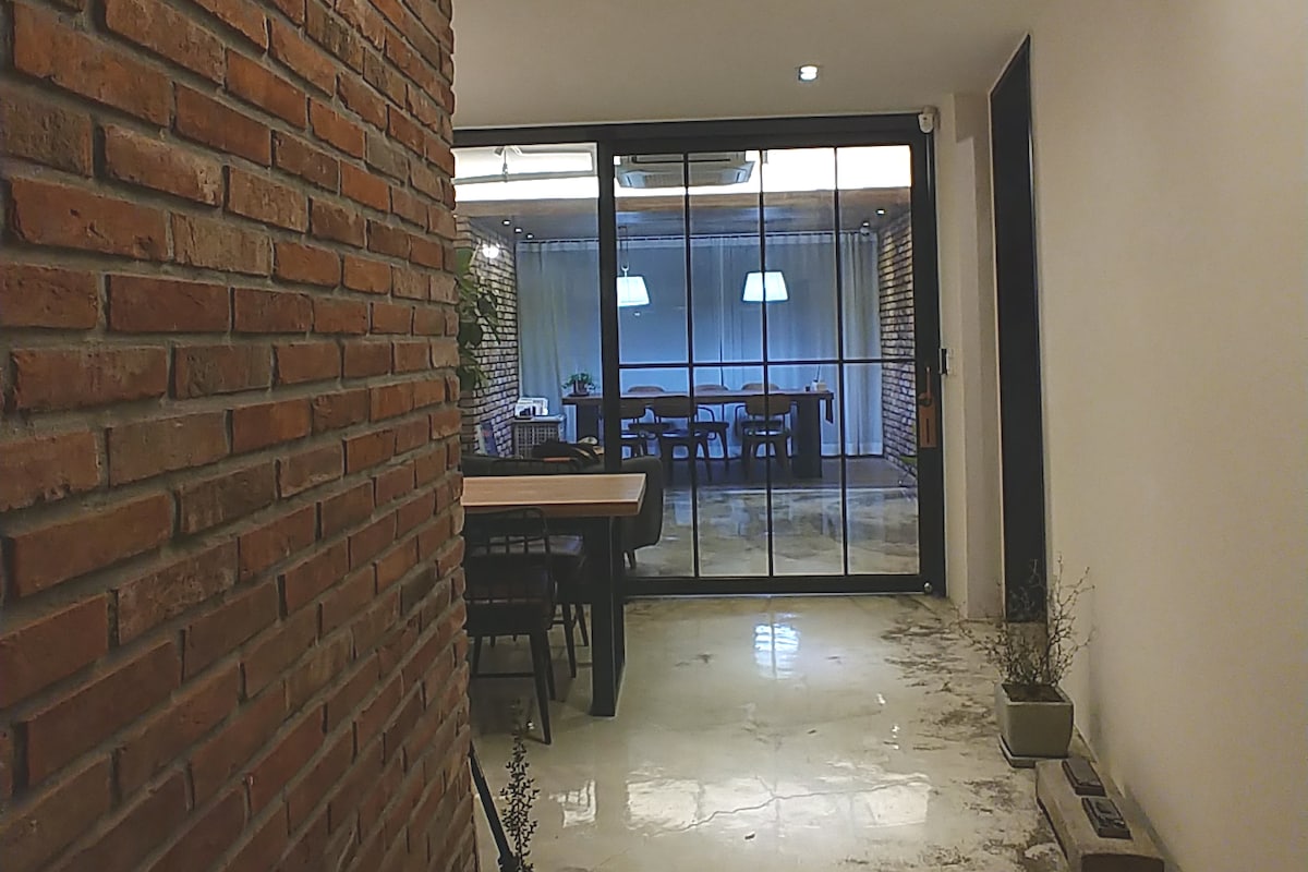 A hallway features a rustic brick wall leading to a spacious dining area filled with natural light. Large glass doors provide a view of the inviting space, where a wooden table surrounded by chairs is placed beneath softly glowing pendant lights.