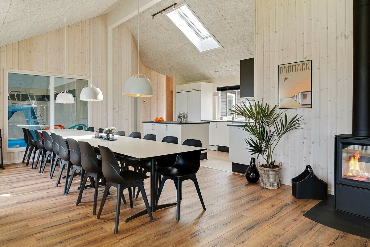 The spacious open-plan living area features a large dining table surrounded by black chairs. A sleek kitchen is visible in the background, complemented by wooden walls and a skylight that enhances natural light. A wood stove creates a cozy focal point in the corner.