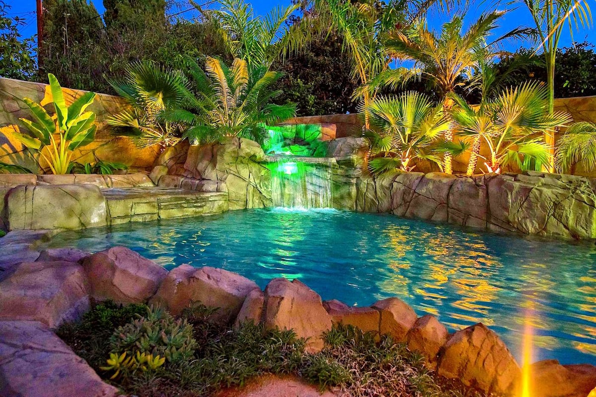 A serene saltwater pool invites relaxation, featuring a rock formation with a waterfall. Lush tropical plants surround the area, illuminated by soft lighting, creating a tranquil atmosphere. The water glows invitingly, reflecting the surrounding greenery and enhancing the peaceful setting.
