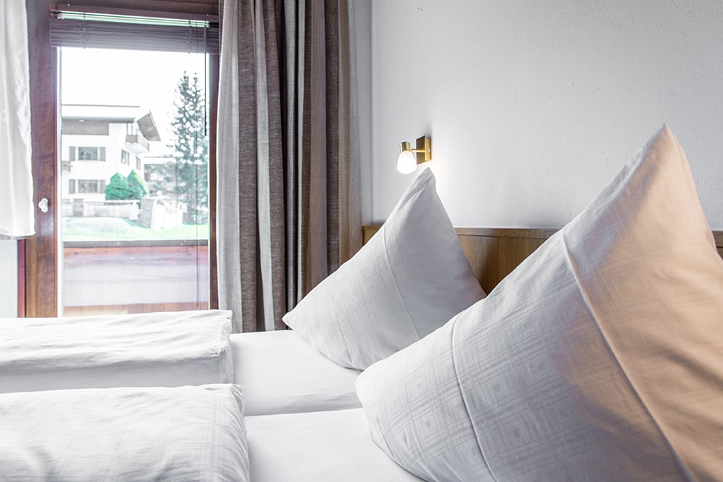 A well-appointed sleeping area features two spacious beds with neatly arranged white linens and decorative pillows. Natural light enters through the large window, which is framed by soft curtains, offering a view of the greenery outside.