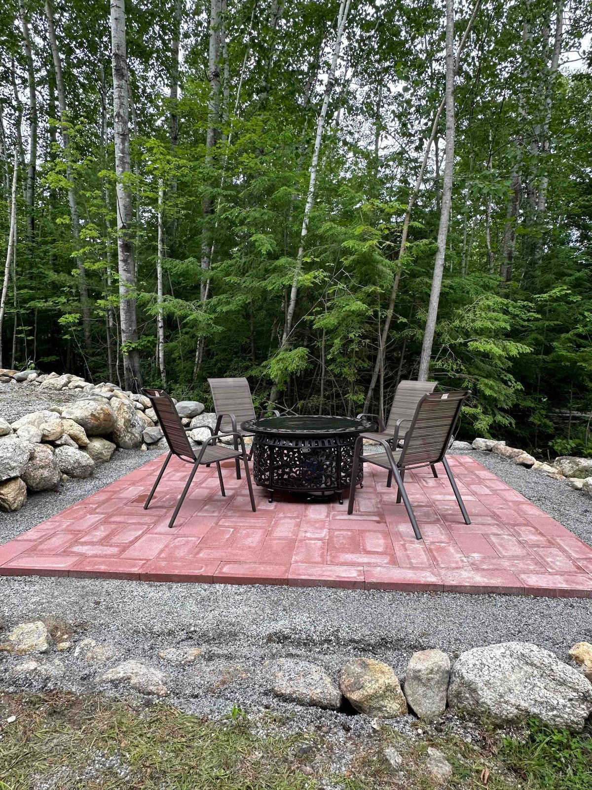 A stone patio area features four metal chairs arranged around a fire pit, set among a natural backdrop of birch trees. Gravel borders the patio, blending into the surrounding landscape.