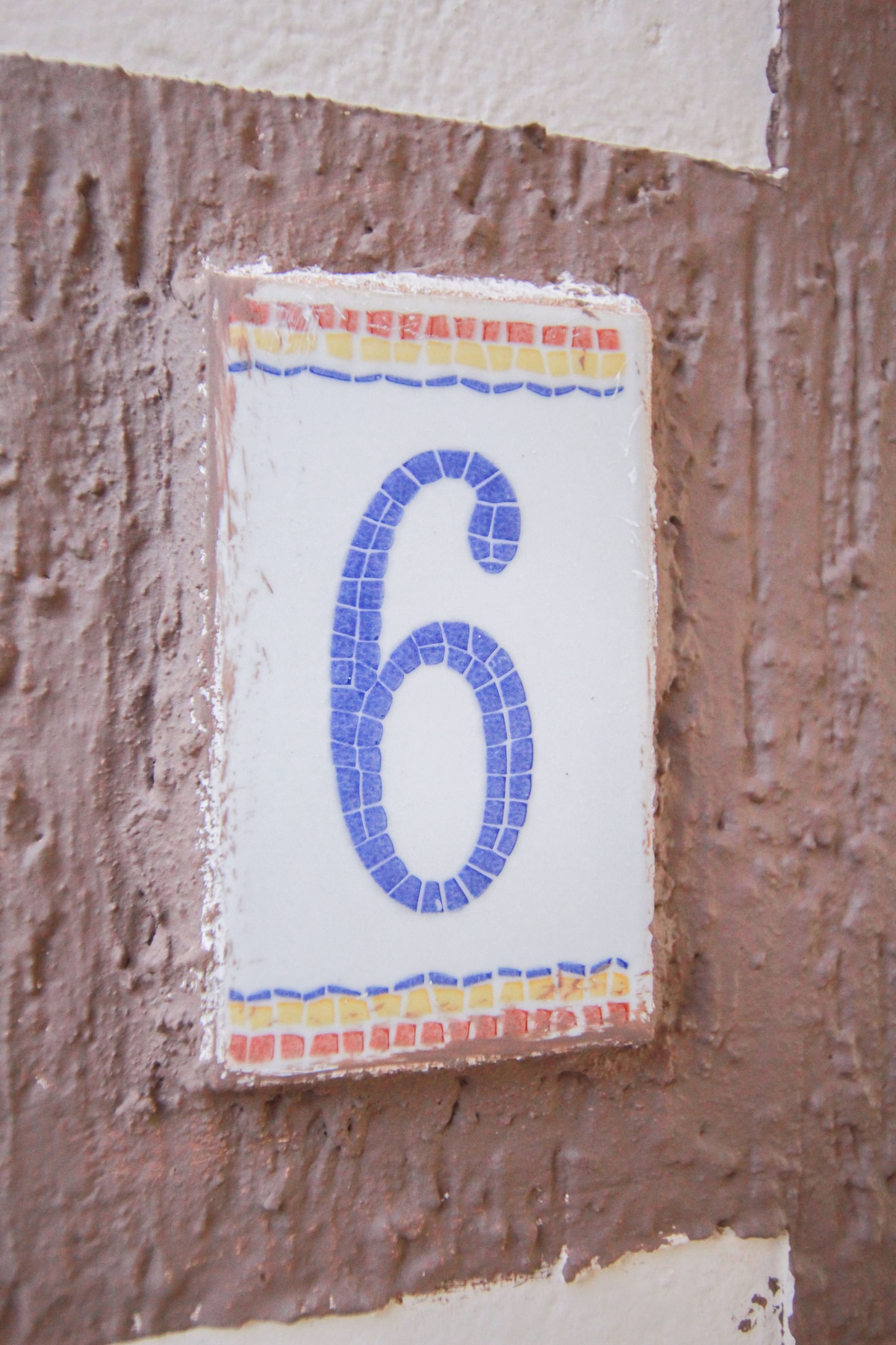 A decorative wall plaque displays the number six in blue mosaic tiles, set against a background of textured brown walls. The edges of the plaque feature a colorful border, adding a vibrant touch to the neutral surroundings.