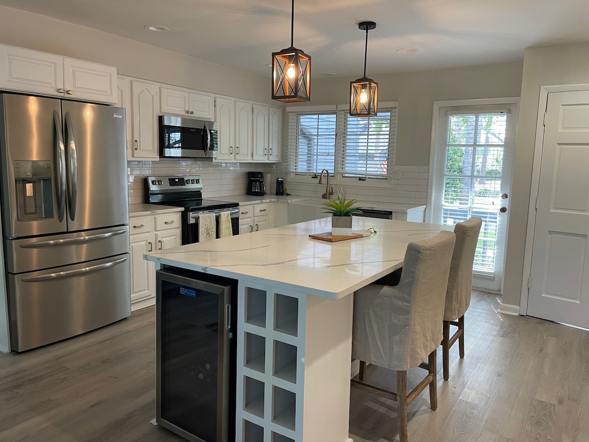 The kitchen features modern stainless steel appliances and an island with seating for three. The white cabinetry complements the light countertops. Natural light is provided through the window and door leading to the outside area, enhancing the spacious feel of the room.