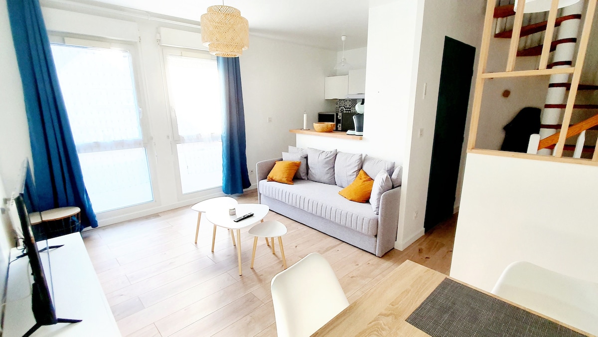 A light-filled living space features a grey sofa adorned with orange cushions, complemented by a minimalistic coffee table. Bright curtains frame large windows, while a staircase leads to an upper level. A dining area is visible with a simple table and chairs.