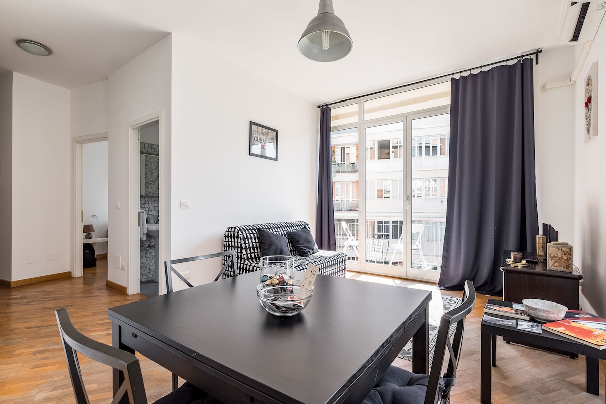 A bright living area features large windows allowing natural light to fill the space. A black and white patterned sofa is positioned across from a dark dining table with a decorative bowl in the center. Dark curtains frame the windows, contributing to the room's modern aesthetic.