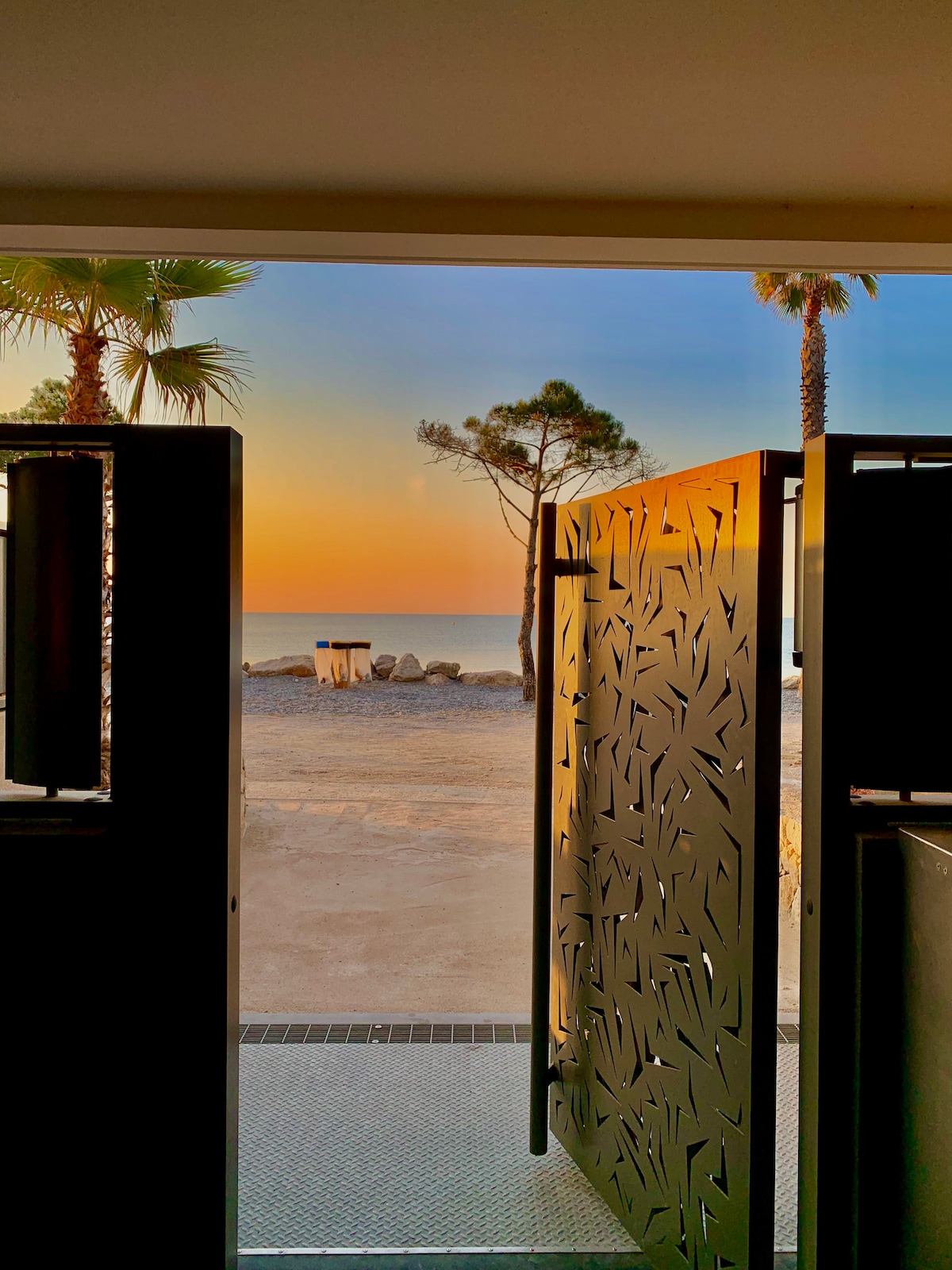 The entrance opens to a serene view of the beach at sunset. Silhouetted palm trees frame the scene, while the sky transitions from warm orange to soft blue. The beach's gentle waves can be seen in the distance.