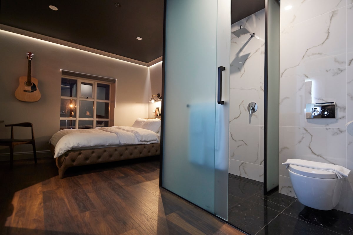 A modern room features a spacious bed with a tufted headboard, accented by wood flooring. A guitar hangs on the wall, and a glass partition separates the bathroom area, which includes a shower and a toilet. Natural light filters through the window.