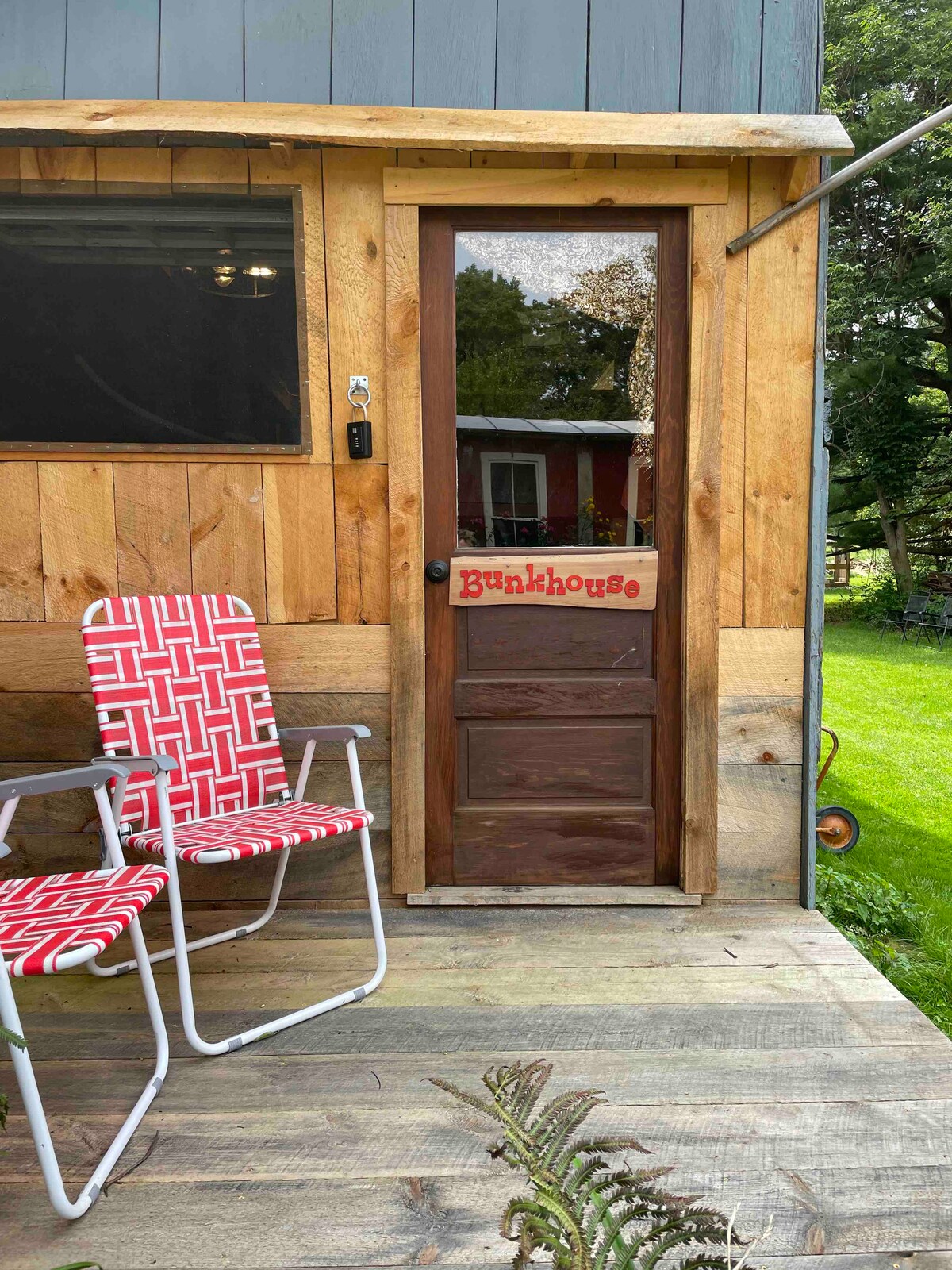 The entrance to the Bunkhouse is framed by rustic wooden siding. A sign labeled 'Bunkhouse' hangs above the wooden door. Two red webbed lawn chairs sit invitingly on the deck, positioned on a wooden floor that leads to a vibrant green lawn.