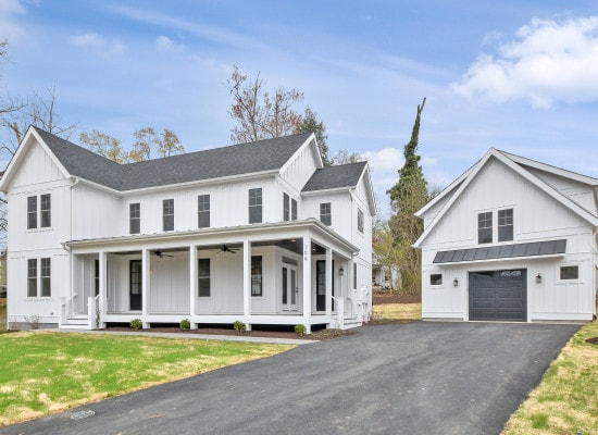 A charming two-story house is displayed alongside a separate carriage house. The exterior is clad in white siding with a gray roof. A covered porch runs the length of the front, supported by columns. A paved driveway provides access to the garage.