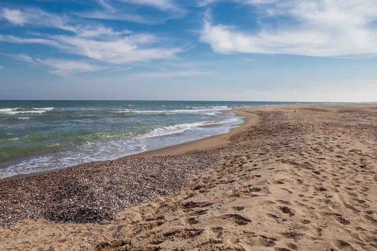 A stretch of sandy beach is seen, meeting the gentle waves of the sea under a clear blue sky. The shore features scattered pebbles, and the horizon reveals the transition between water and sky.