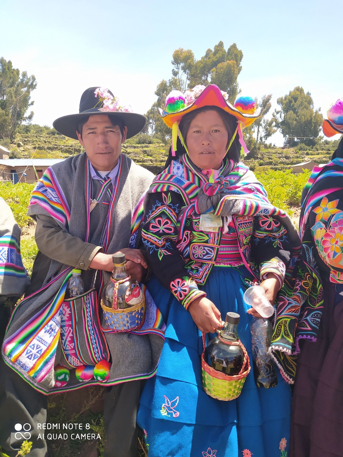 Two individuals are dressed in traditional Andean clothing, showcasing vibrant patterns and colors. One holds a large jug, while the other carries a woven basket. The backdrop features greenery and a clear blue sky, highlighting the cultural attire and accessories.