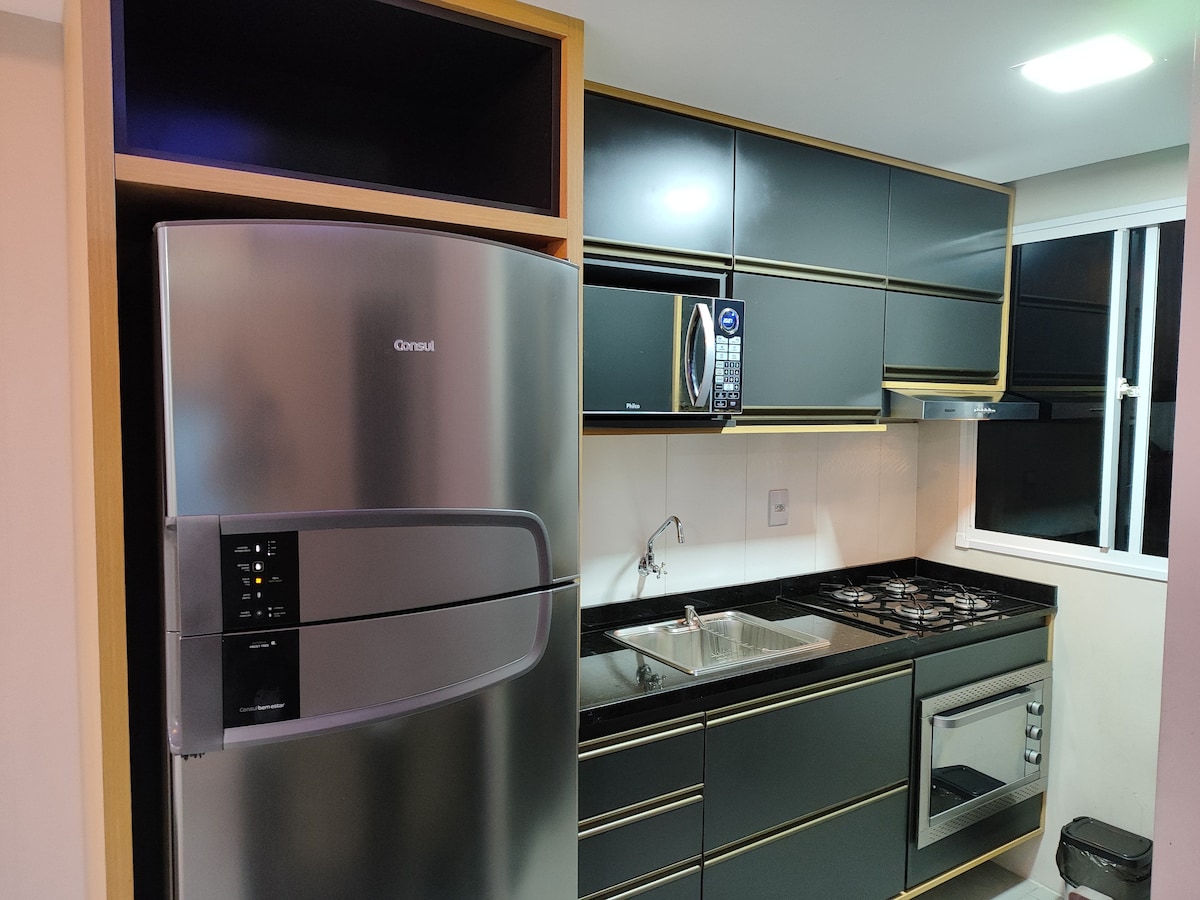 A modern kitchen is shown, featuring sleek black cabinetry and a stainless steel refrigerator. A built-in microwave and a gas stove are visible, accompanied by a stainless steel sink. Natural light enters through a window, illuminating the countertop and appliances.