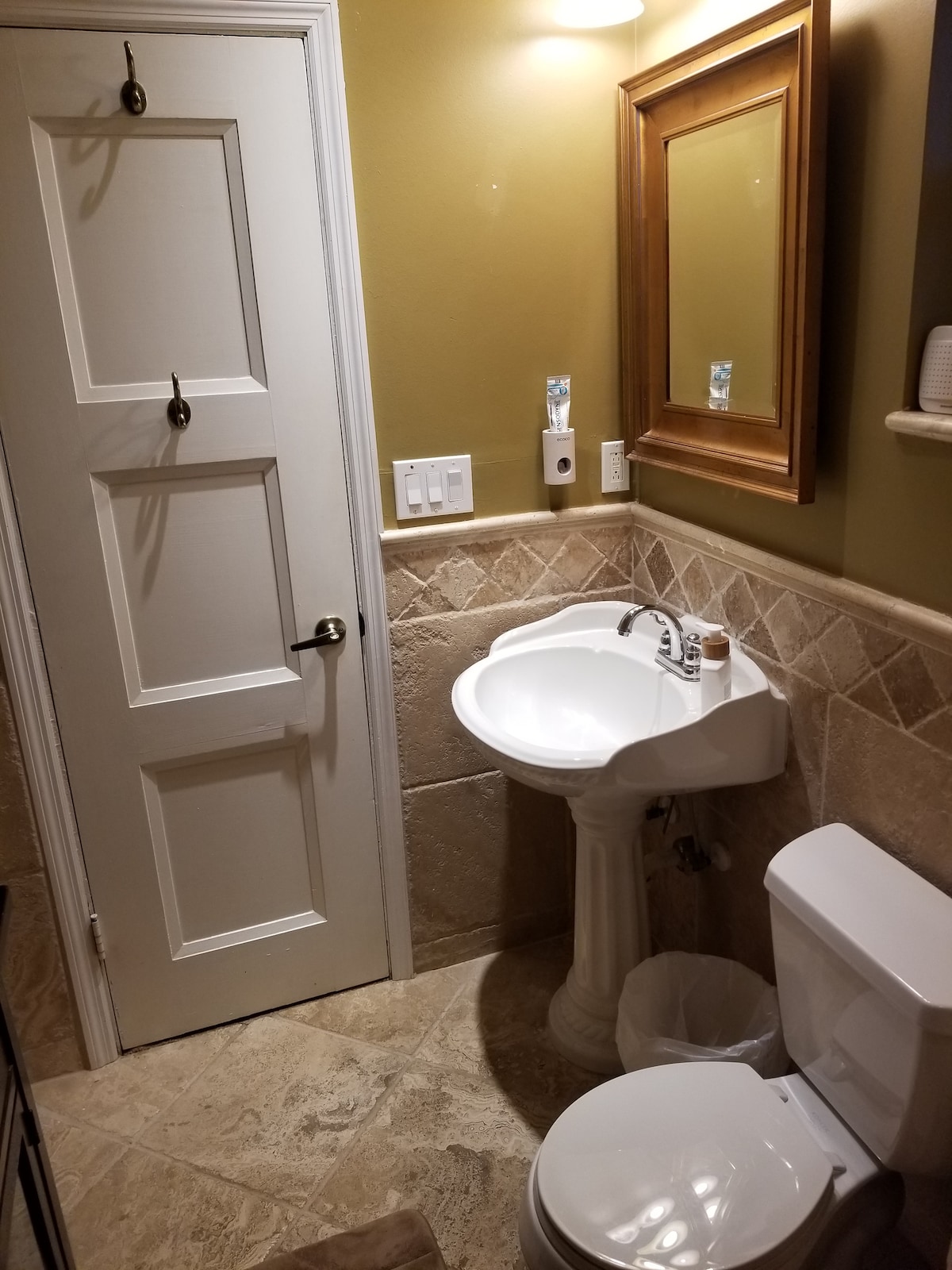 A private bathroom features neutral-toned walls and natural stone tile flooring. A small pedestal sink is situated next to a sleek toilet. A mirror with a decorative frame is mounted above the sink. The space is illuminated by warm overhead lighting.